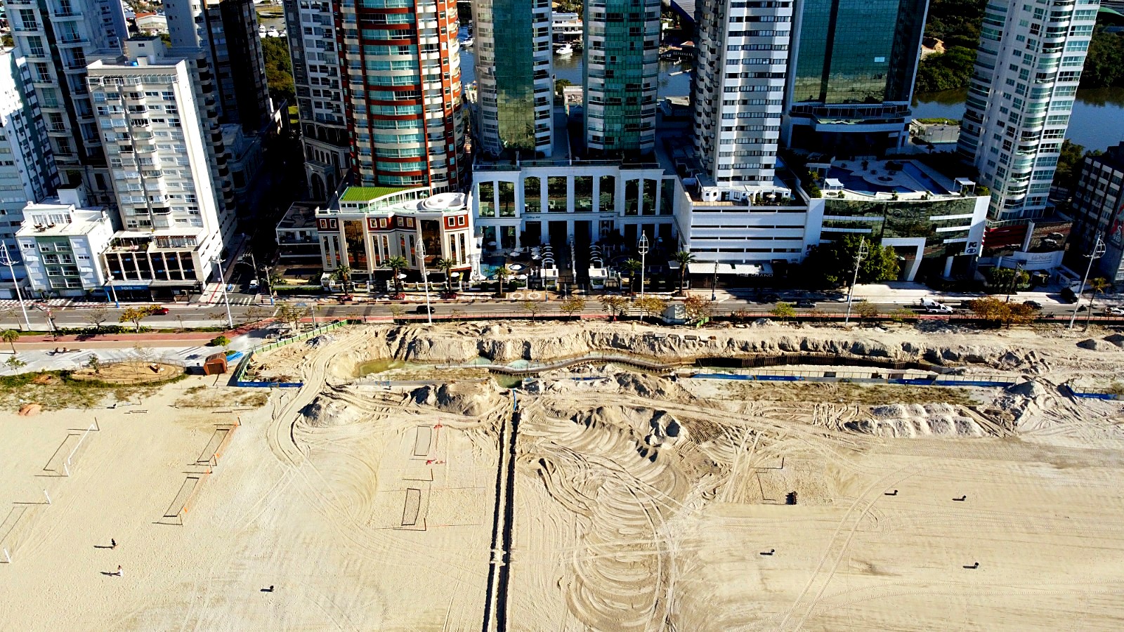 Vista aérea de cima da orla de Balneário Camboriú, com os prédios e a Avenida Atlântica à esquerda e, entre a avenida e a faixa de areia, uma faixa larga de terreno escavado com montes de areia, marcas de máquina e uma vala com água. Na areia aparecem quadras demarcadas e algumas pessoas.
