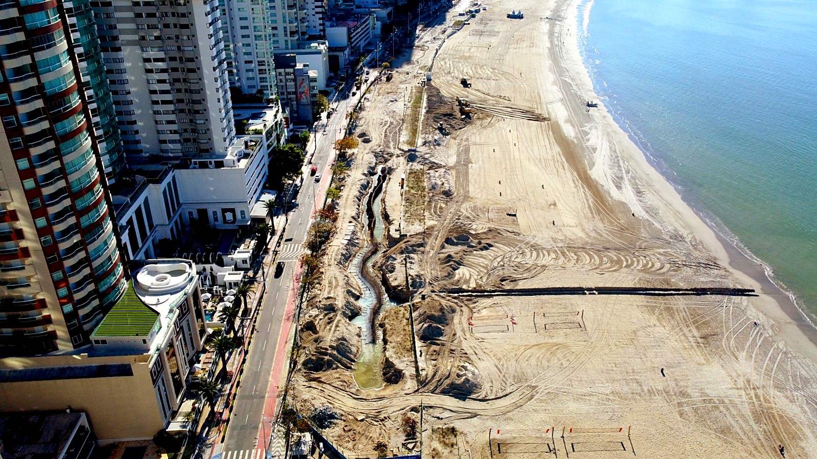 Vista aérea inclinada da praia de Balneário Camboriú olhando para o norte, com o mar à direita, a faixa de areia larga no centro e, junto à Avenida Atlântica, a área escavada da obra com um canal de água serpenteando entre montes de terra e máquinas trabalhando.